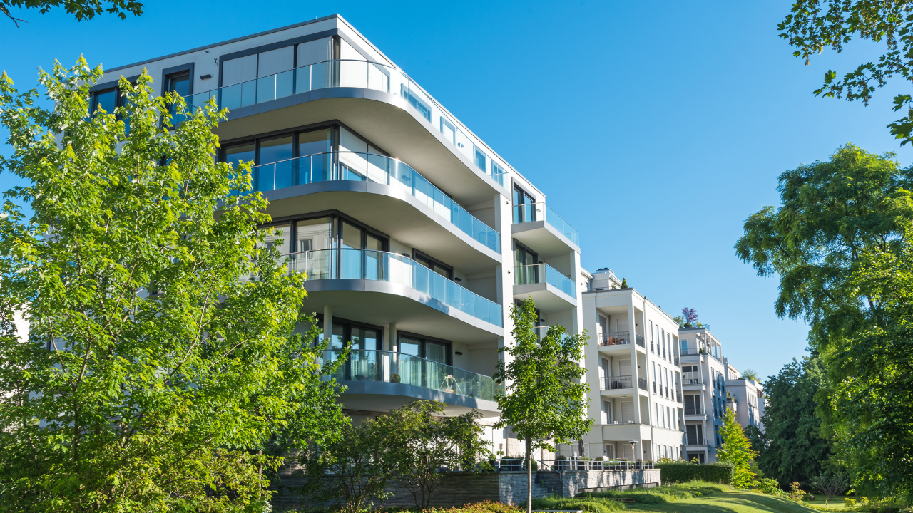 Modern apartment buildings with curved glass balconies surrounded by leafy trees under a clear blue sky.