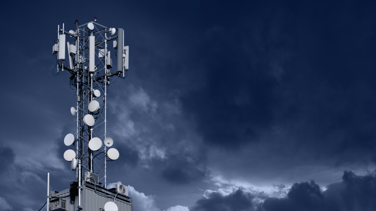 Telecommunications tower equipped with cellular antennas and satellite dishes beneath a dark, cloudy sky.